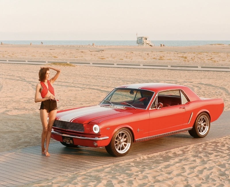 A woman in a red top and black shorts stands barefoot on a boardwalk at the beach, posing next to a classic red Ford Mustang coupe under warm sunset light, with the ocean and a lifeguard tower visible in the background